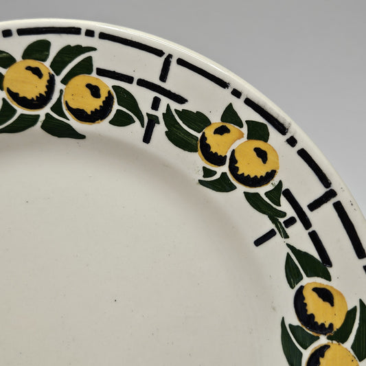 De couleur beige très pâle, comme beaucoup de faïences, cette assiette est entourée de fruits ronds jaunes avec des ombres noires prononcées sur le bas de chaque fruit en séquence de 3 - 2 - 3 - 2 . Chaque séquence est entourée de feuilles vertes et le tout est posé sur un fin quadrillé noir. Les couleurs sont chatoyantes.
Aucune restauration