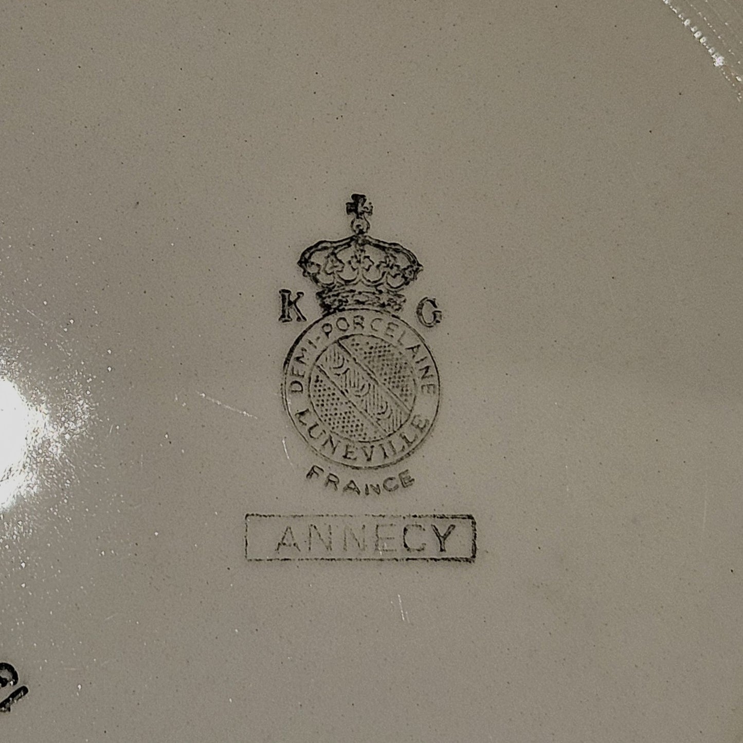 Vue du dos de l'assiette en demi-porcelaine Keller & Guerin Lunéville modèle Annecy, montrant le poinçon manufacture : couronne royale surmontant le médaillon K & G / Demi-Porcelaine / Lunéville / France, avec le nom du modèle Annecy dans un cartouche rectangulaire, sur fond beige.