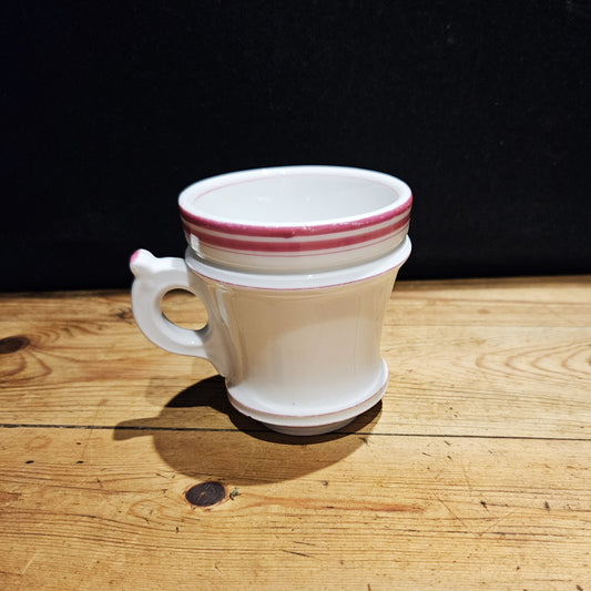 Tasse à brûlot ancienne en faïence blanche avec une petite anse ronde, ornée de trois rayures roses sur le bord supérieur, posée sur une surface en bois brut, fond noir.