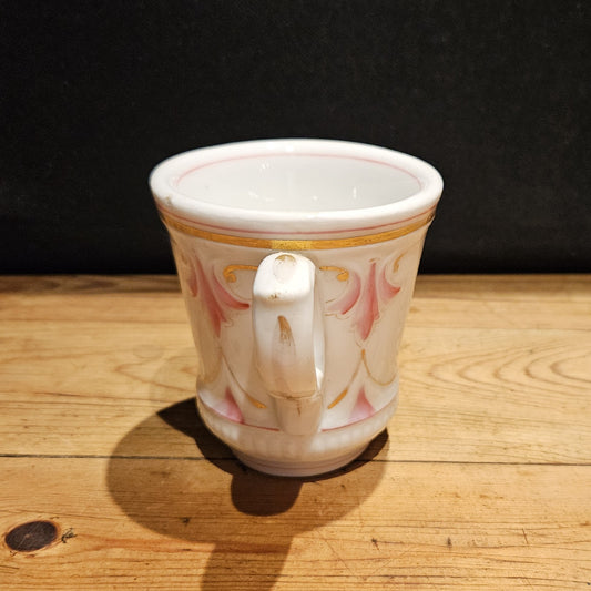 Vue arrière de la tasse à brûlot en faïence blanche, montrant l'anse ronde et le décor en relief de volutes roses et dorées sur tout le pourtour, fond noir.