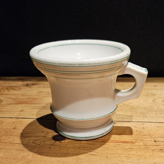 Vue de trois quarts de la tasse à brûlot en faïence blanche, montrant la forme tronconique évasée, les filets verts et or et l'anse rectangulaire à ouverture ronde, posée sur une table en bois, fond noir.