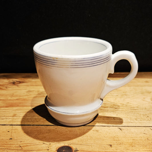 Tasse à brûlot ancienne en faïence blanche avec fins filets bleus, anse ronde à droite, pied bombé, posée sur une table en bois brut, fond noir.