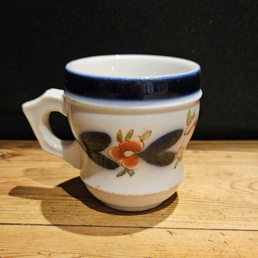 Tasse à brûlot ancienne en faïence blanche, avec large bandeau bleu cobalt sur le bord supérieur, décor floral peint à la main (fleurs orangées et feuilles sombres), anse à gauche, posée sur une table en bois, fond noir.