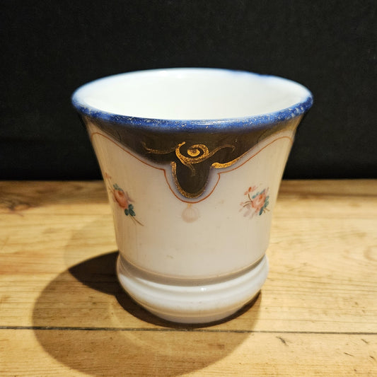 Vue de face de la tasse à brûlot en faïence blanche, montrant le bandeau bleu cobalt, les volutes dorées et les petites roses peintes à la main, sans anse visible, posée sur une table en bois brut, fond noir.