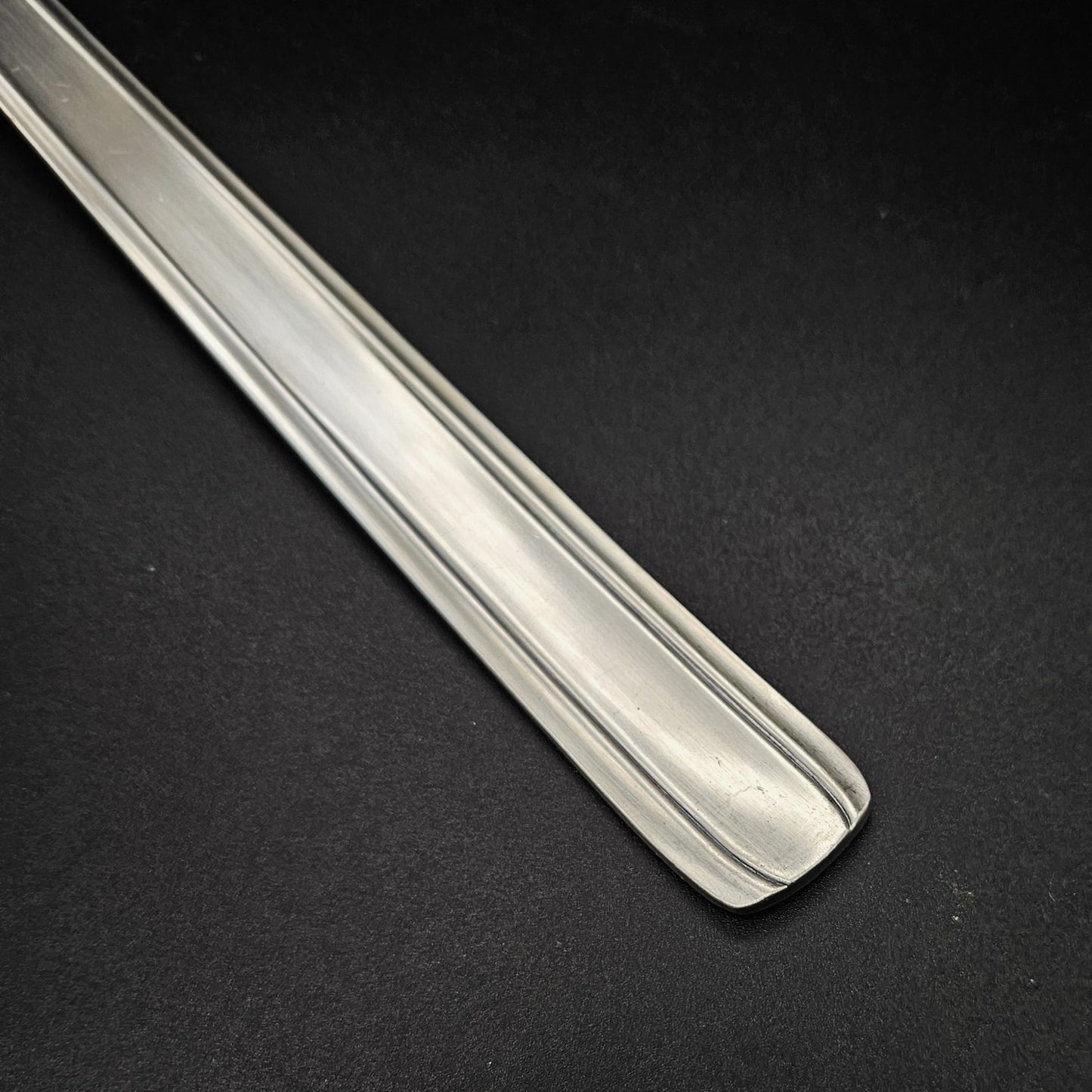 Gros plan sur l'extrémité carrée légèrement arrondie du manche de la louche à potage en métal argenté Moderniste, montrant le double filet latéral fin courant sur toute la longueur du manche plat et rectiligne, sur fond noir.