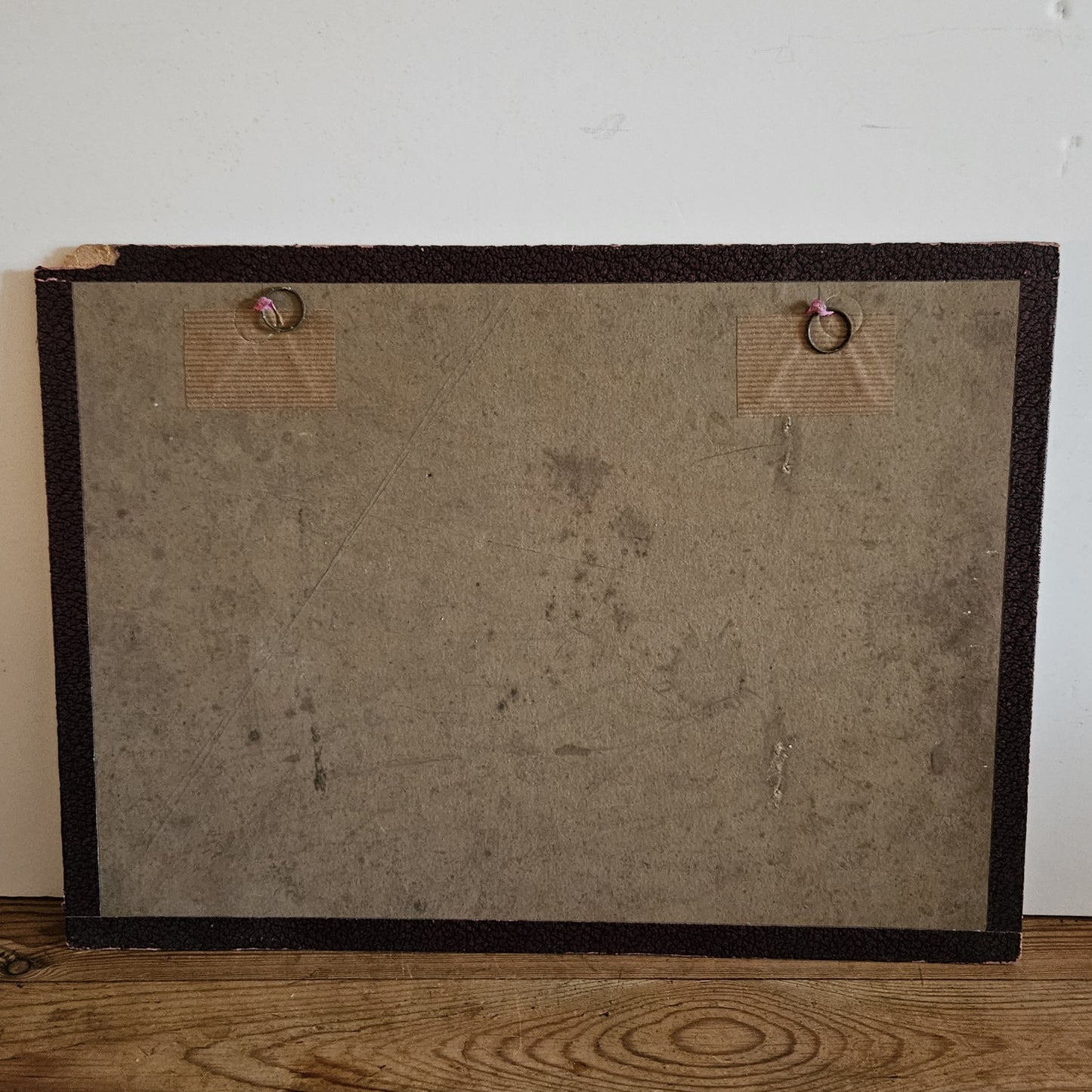 Vue du dos du cadre de l'aquarelle de Paul Friesé, montrant un fond en carton brun avec deux anneaux de suspension fixés par des étiquettes en carton, posé sur un parquet en bois brut.