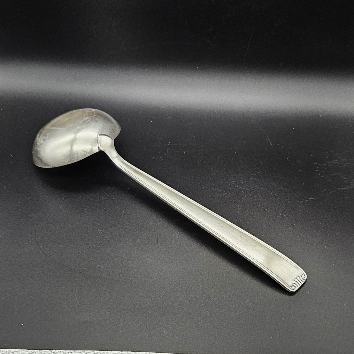 Louche à potage en métal argenté Art Déco, 31 cm, vue de dos sur fond noir, montrant le dos de la cuillère ronde et le manche rectangulaire à double filet latéral avec extrémité en éventail à cannelures.