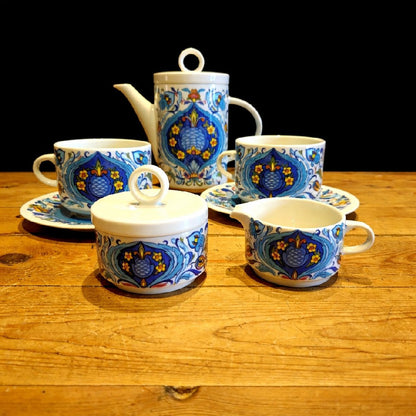 Service à thé en porcelaine décorative composé de deux tasses assorties, ornées de motifs floraux colorés en bleu, jaune et rouge. Les motifs, symétriques et détaillés, évoquent un style artisanal méditerranéen. Les tasses présentent une finition brillante et des formes élégantes légèrement arrondies. Elles reposent sur une surface en bois clair au grain visible, qui contraste avec un fond sombre et met en valeur les couleurs vives et les détails du décor. 