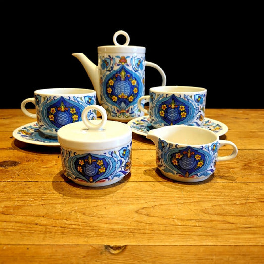 Service à thé en porcelaine décorative composé de deux tasses assorties, ornées de motifs floraux colorés en bleu, jaune et rouge. Les motifs, symétriques et détaillés, évoquent un style artisanal méditerranéen. Les tasses présentent une finition brillante et des formes élégantes légèrement arrondies. Elles reposent sur une surface en bois clair au grain visible, qui contraste avec un fond sombre et met en valeur les couleurs vives et les détails du décor. 