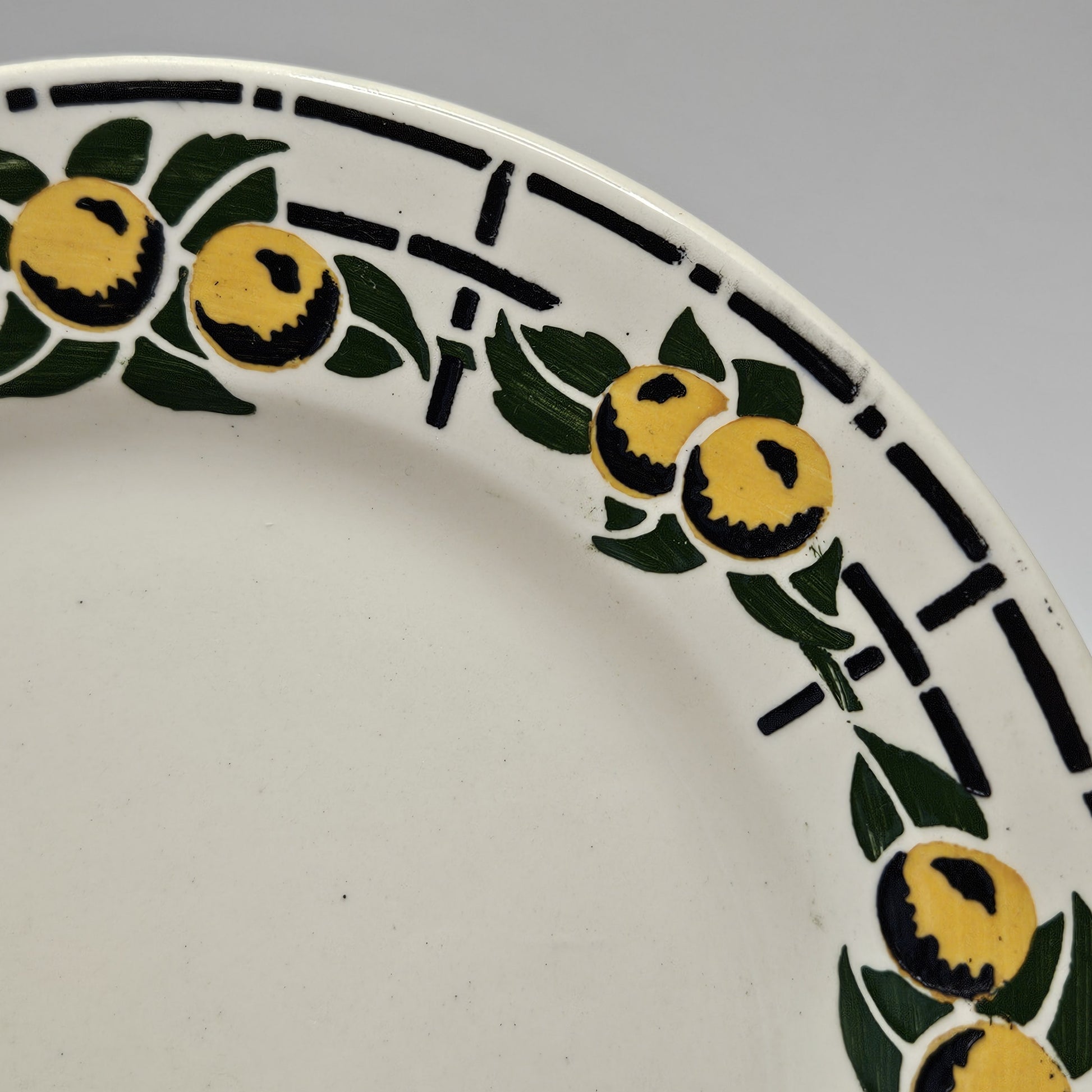 De couleur beige très pâle, comme beaucoup de faïences, cette assiette est entourée de fruits ronds jaunes avec des ombres noires prononcées sur le bas de chaque fruit en séquence de 3 - 2 - 3 - 2 . Chaque séquence est entourée de feuilles vertes et le tout est posé sur un fin quadrillé noir. Les couleurs sont chatoyantes.
Aucune restauration