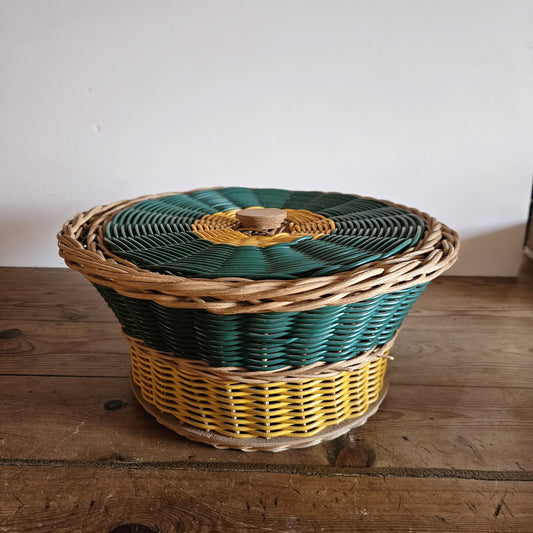 Vue de face de la panière ronde à couvercle en osier tressé tricolore vert, jaune et naturel, avec bouton de préhension en bois tourné, posée sur une table en bois brut, fond blanc.
