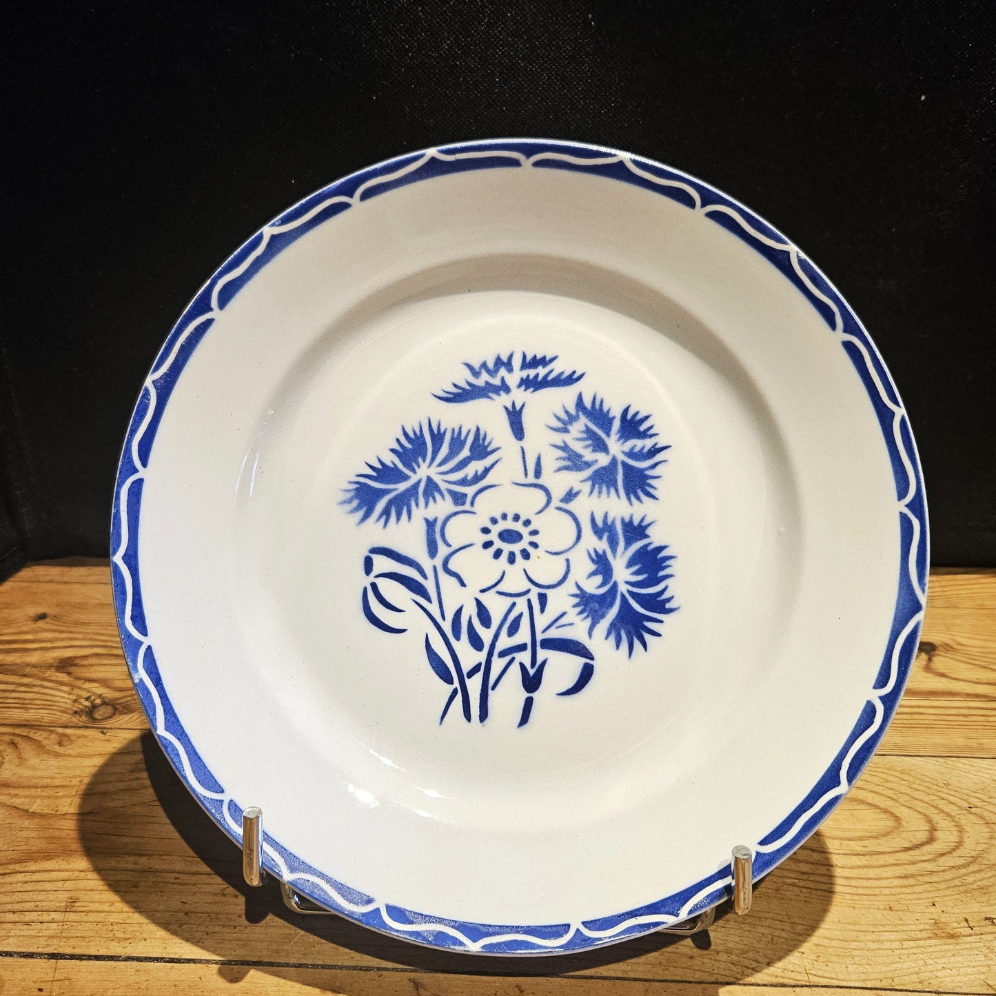 Vue générale de l'assiette plate en demi-porcelaine Keller & Guerin Lunéville modèle Annecy, Ø 23 cm, montrant le décor complet : bouquet central de bleuets et anémone au pochoir bleu cobalt et bordure à vagues entrelacées réservées sur fond bleu, posée sur un présentoir en métal chromé sur fond bois.