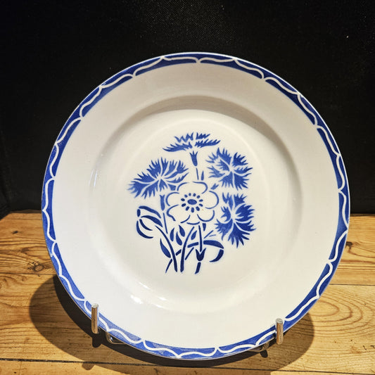 Vue générale de l'assiette plate en demi-porcelaine Keller & Guerin Lunéville modèle Annecy, Ø 23 cm, montrant le décor complet : bouquet central de bleuets et anémone au pochoir bleu cobalt et bordure à vagues entrelacées réservées sur fond bleu, posée sur un présentoir en métal chromé sur fond bois.