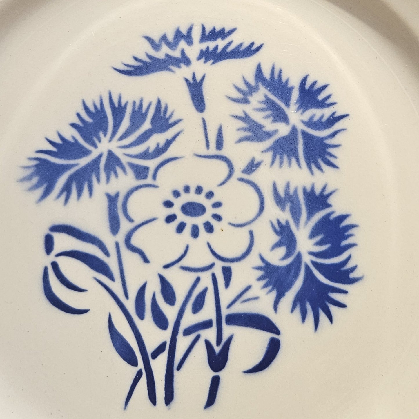 Détail du décor central de l'assiette en demi-porcelaine Keller & Guerin Lunéville modèle Annecy, montrant le bouquet de bleuets aux pétales finement découpés et d'une anémone à cinq pétales avec cœur à pistils, tiges et feuilles stylisées, au pochoir bleu cobalt sur fond blanc.