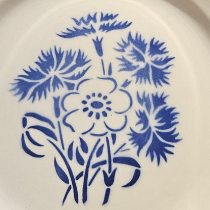 Détail du décor central de l'assiette en demi-porcelaine Keller & Guerin Lunéville modèle Annecy, montrant le bouquet de bleuets aux pétales finement découpés et d'une anémone à cinq pétales avec cœur à pistils, tiges et feuilles stylisées, au pochoir bleu cobalt sur fond blanc.