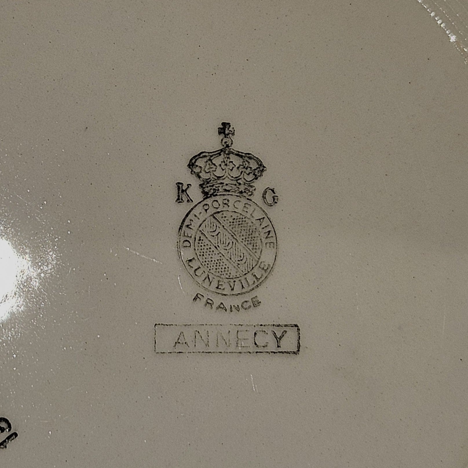 Vue du dos de l'assiette en demi-porcelaine Keller & Guerin Lunéville modèle Annecy, montrant le poinçon manufacture : couronne royale surmontant le médaillon K & G / Demi-Porcelaine / Lunéville / France, avec le nom du modèle Annecy dans un cartouche rectangulaire, sur fond beige.