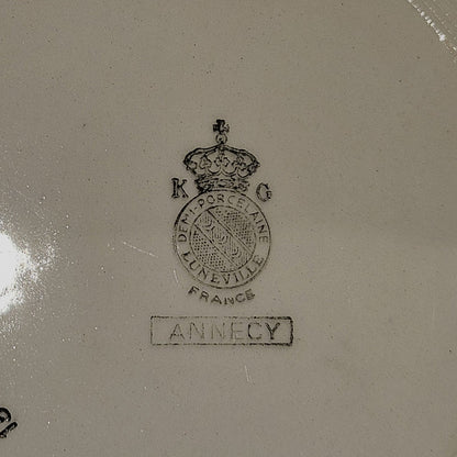 Vue du dos de l'assiette en demi-porcelaine Keller & Guerin Lunéville modèle Annecy, montrant le poinçon manufacture : couronne royale surmontant le médaillon K & G / Demi-Porcelaine / Lunéville / France, avec le nom du modèle Annecy dans un cartouche rectangulaire, sur fond beige.