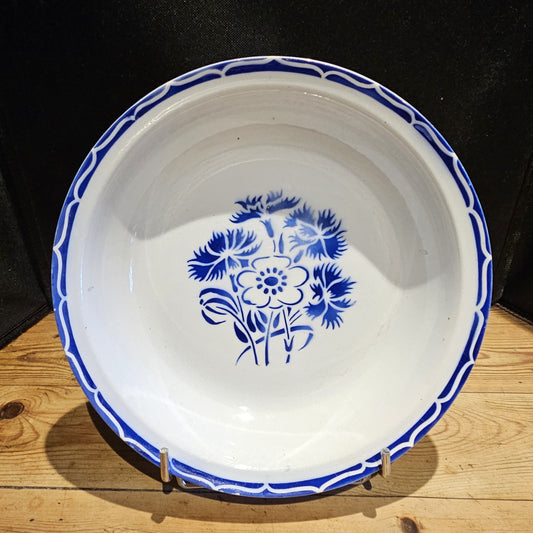 Plat creux en porcelaine opaque Keller & Guerin Lunéville modèle Annecy, Ø 25 cm, vue de face sur fond noir, montrant le décor complet : bouquet central de bleuets et anémone au pochoir bleu cobalt et bordure à vagues entrelacées réservées sur fond bleu cobalt, posé sur un présentoir en métal chromé sur fond bois.