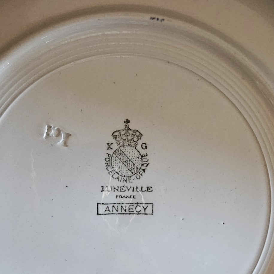 Gros plan sur le poinçon du plat creux Keller & Guerin Lunéville modèle Annecy : couronne royale surmontant le médaillon K & G / Porcelaine Opaque / Lunéville / France, avec le nom du modèle Annecy dans un cartouche rectangulaire et le numéro de moule 121 en relief, sur fond blanc ivoire.