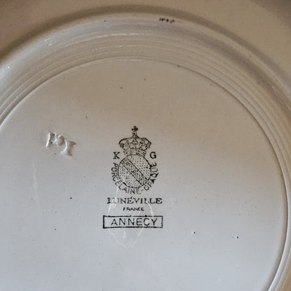 Gros plan sur le poinçon du plat creux Keller & Guerin Lunéville modèle Annecy : couronne royale surmontant le médaillon K & G / Porcelaine Opaque / Lunéville / France, avec le nom du modèle Annecy dans un cartouche rectangulaire et le numéro de moule 121 en relief, sur fond blanc ivoire.