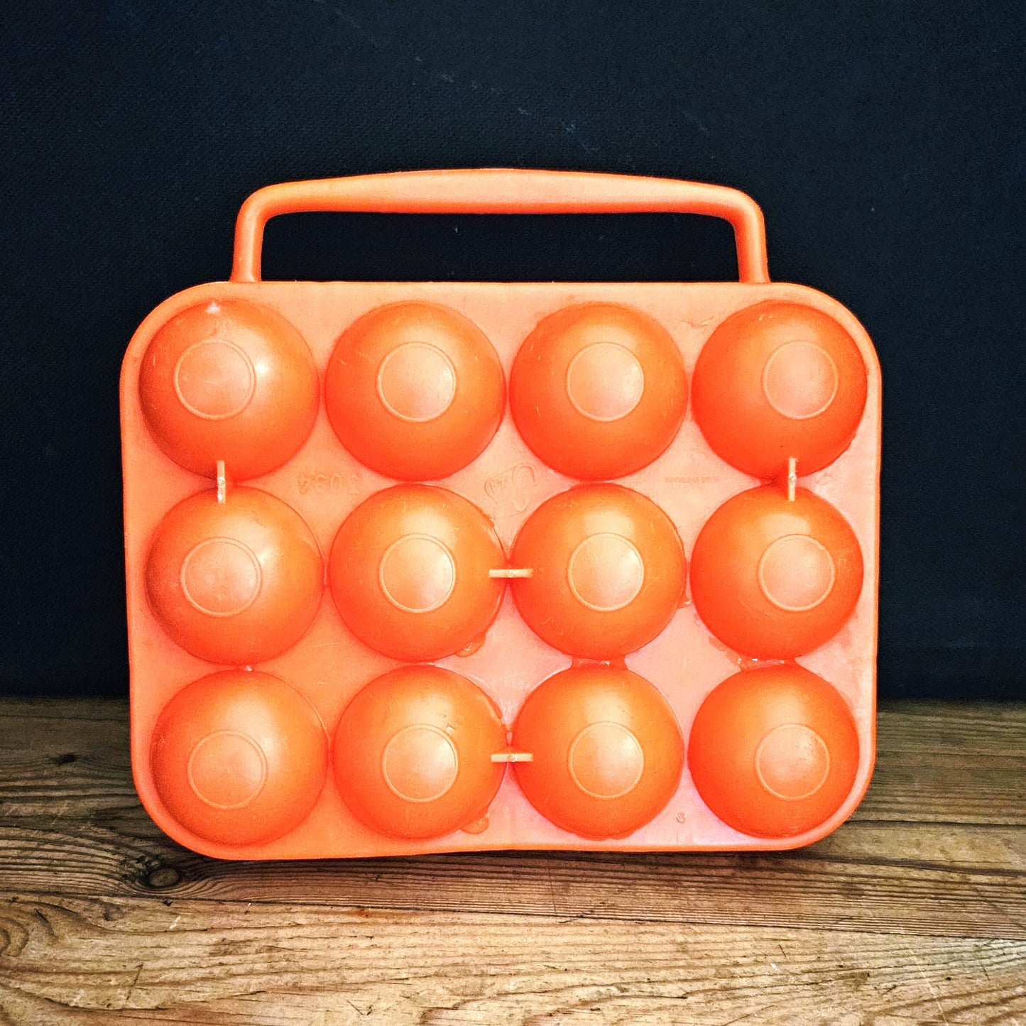 Boîte à œufs vintage en plastique orange vif Made in France, années 1970, vue fermée de face sur fond noir, montrant les 12 alvéoles bombées en deux rangées de 4 x 3 et la poignée centrale intégrée, posée sur une table en bois brut.