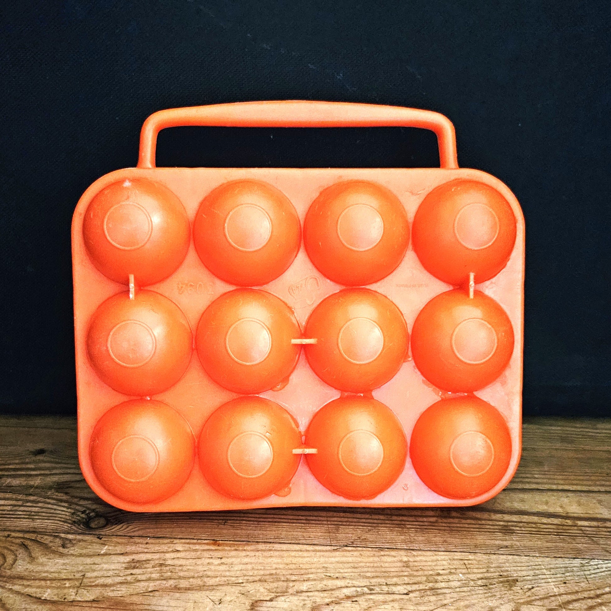 Boîte à œufs vintage en plastique orange vif Made in France, années 1970, vue fermée de face sur fond noir, montrant les 12 alvéoles bombées en deux rangées de 4 x 3 et la poignée centrale intégrée, posée sur une table en bois brut.