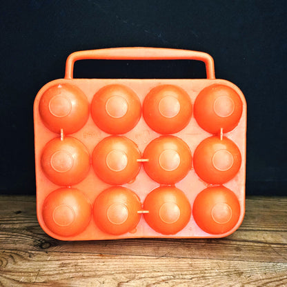 Boîte à œufs vintage en plastique orange vif Made in France, années 1970, vue fermée de face sur fond noir, montrant les 12 alvéoles bombées en deux rangées de 4 x 3 et la poignée centrale intégrée, posée sur une table en bois brut.