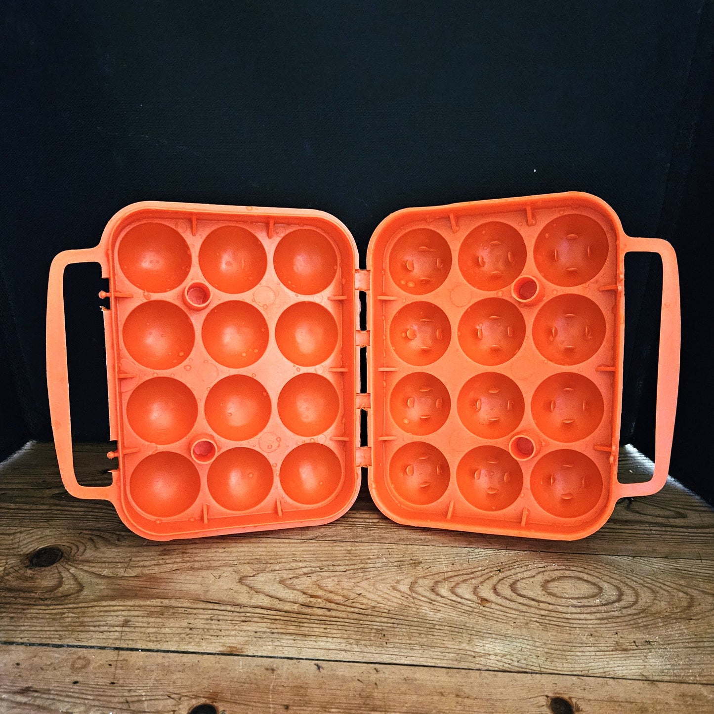 Boîte à œufs vintage en plastique orange vif Made in France, années 1970, vue ouverte sur fond noir, montrant les deux demi-coques symétriques de 3 x 4 alvéoles avec charnière centrale et poignées latérales intégrées, posée sur une table en bois brut.