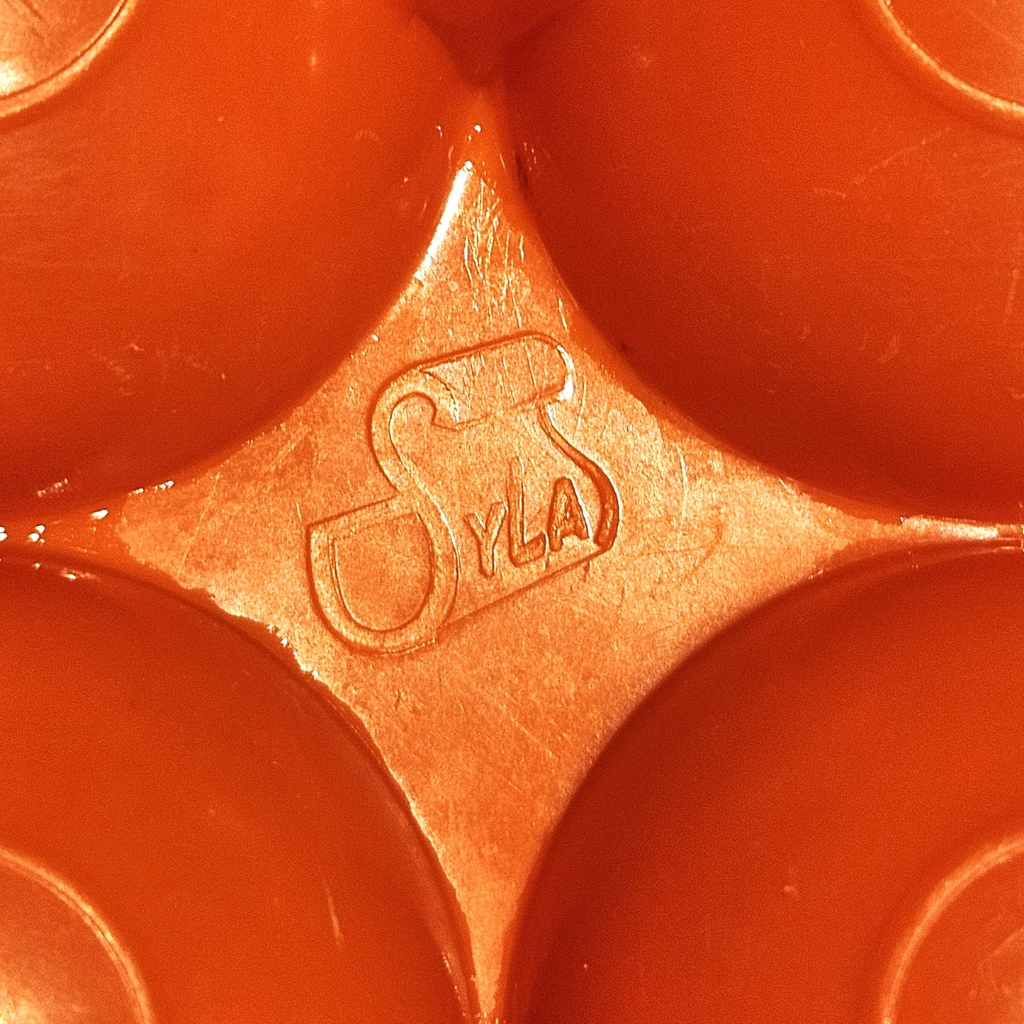 Gros plan sur le marquage Syla moulé dans le plastique orange vif de la boîte à œufs vintage, visible entre les alvéoles bombées, sur fond orange.