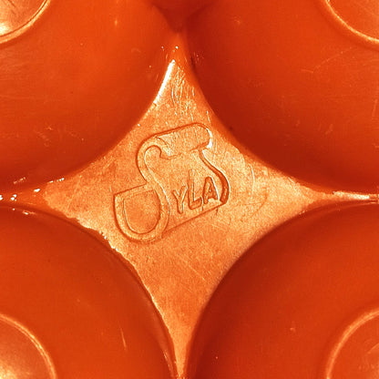 Gros plan sur le marquage Syla moulé dans le plastique orange vif de la boîte à œufs vintage, visible entre les alvéoles bombées, sur fond orange.