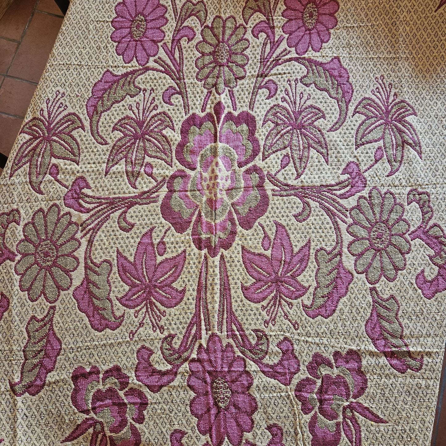 Vue générale du jeté de lit en jacquard double face années 1950-1960, face envers fond crème/écru, montrant le décor floral Arts & Crafts en rose framboise avec bordure à losanges géométriques entrelacés, posé à plat sur carrelage tomette rouge.