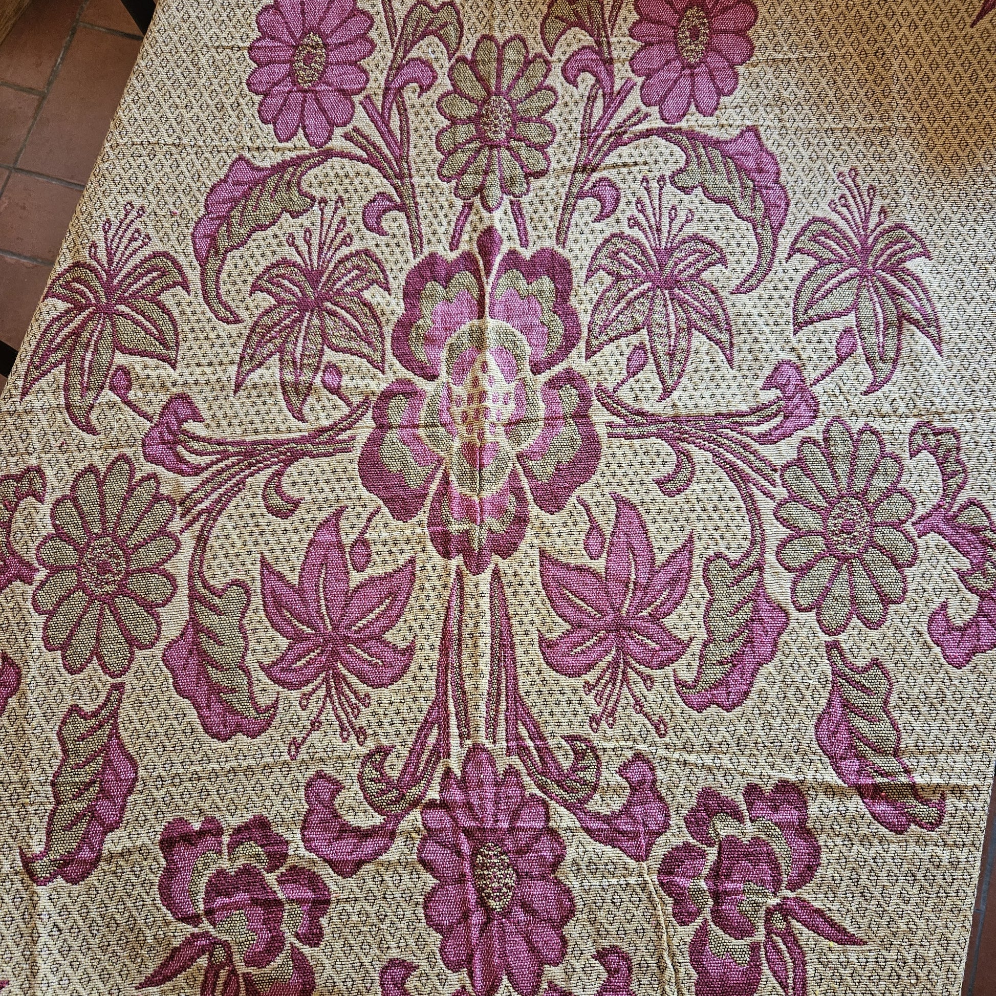 Vue générale du jeté de lit en jacquard double face années 1950-1960, face envers fond crème/écru, montrant le décor floral Arts & Crafts en rose framboise avec bordure à losanges géométriques entrelacés, posé à plat sur carrelage tomette rouge.