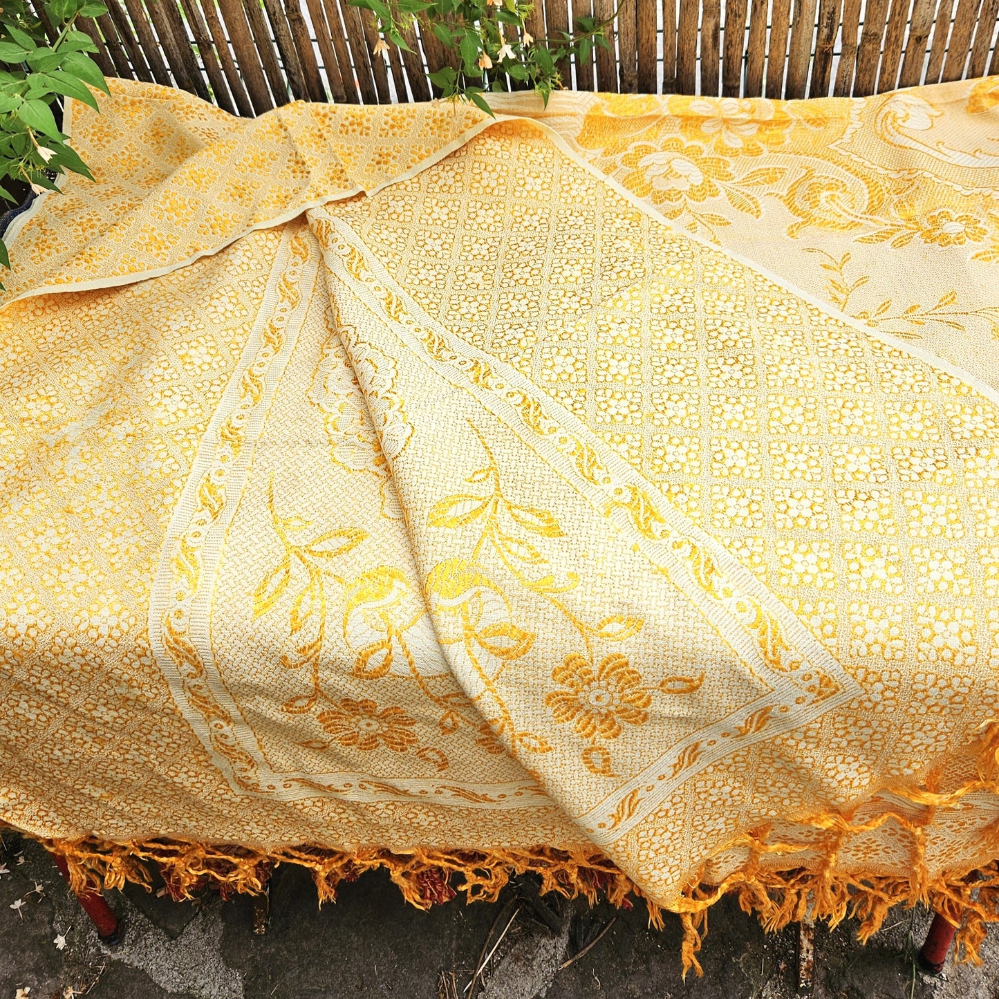 Jeté de canapé en jacquard double face années 1950-1960, camaïeu or et crème, posé sur un banc de jardin en bois avec franges torsadées or/jaune visibles sur le petit côté, fond de bambouseraie et végétation.