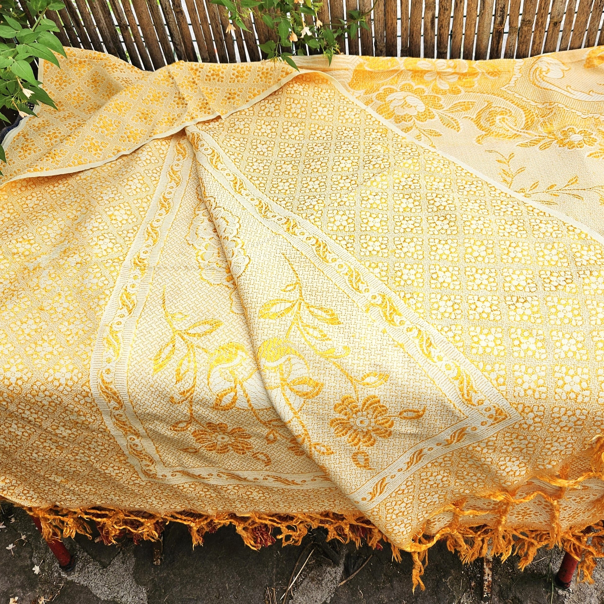 Jeté de canapé en jacquard double face années 1950-1960, camaïeu or et crème, posé sur un banc de jardin en bois avec franges torsadées or/jaune visibles sur le petit côté, fond de bambouseraie et végétation.