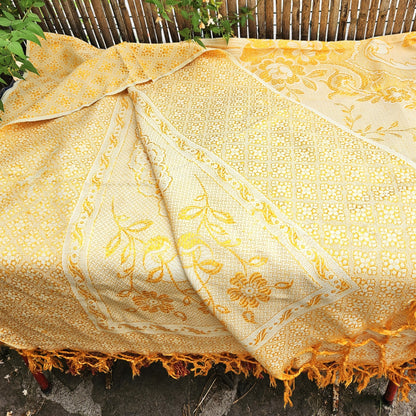 Jeté de canapé en jacquard double face années 1950-1960, camaïeu or et crème, posé sur un banc de jardin en bois avec franges torsadées or/jaune visibles sur le petit côté, fond de bambouseraie et végétation.