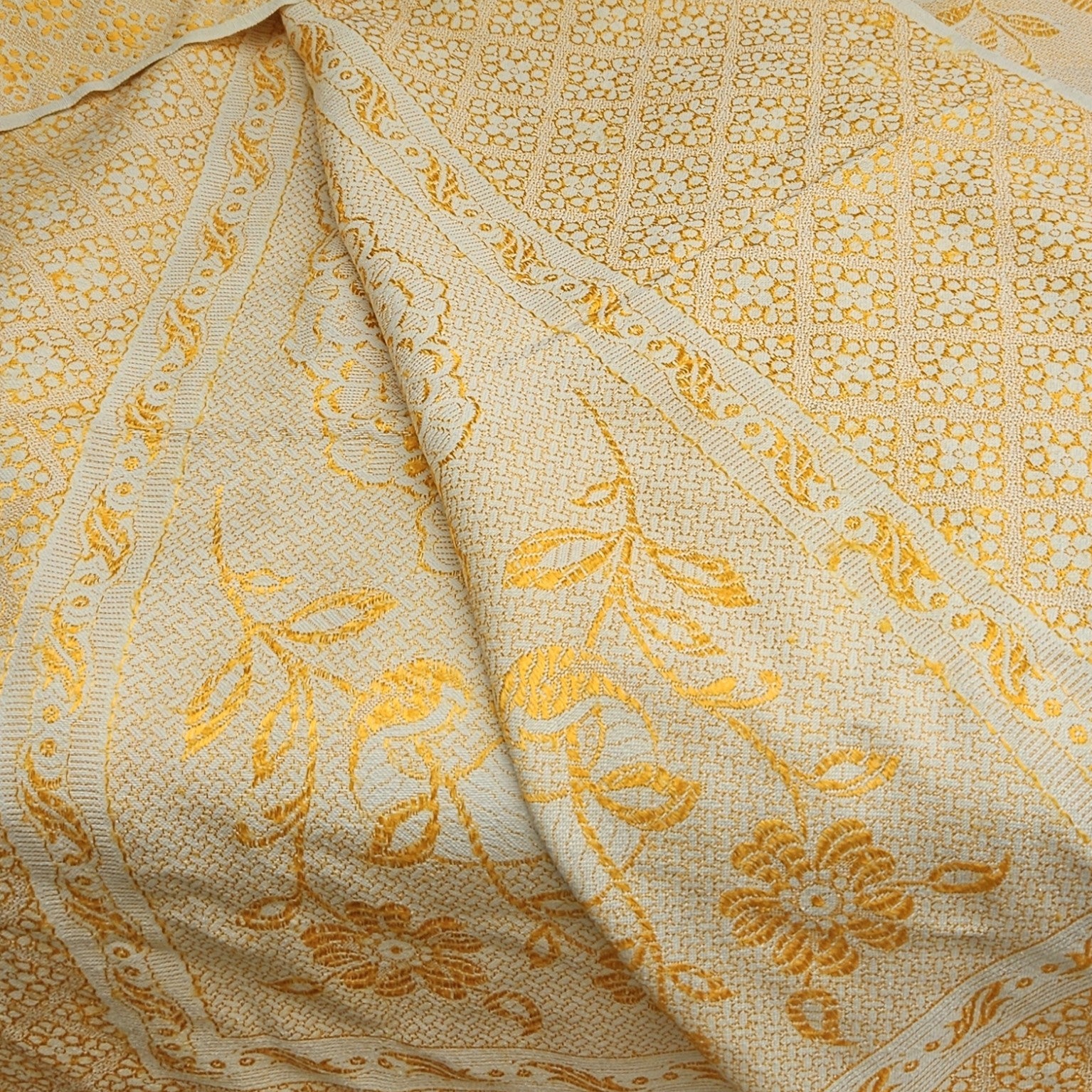 Détail de la frise florale à feuillages et rinceaux du jeté de canapé en jacquard double face années 1950-1960, montrant les deux faces du tissu en repli — fond crème/or chaud côté endroit et fond or/jaune intense côté envers — avec résille de petits motifs géométriques en nid d'abeilles visible sur le fond.