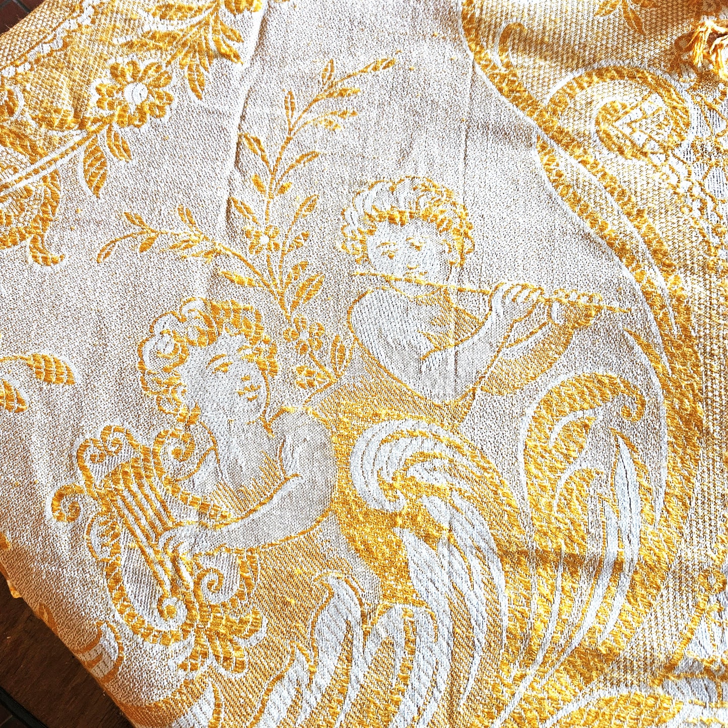 Détail d'un coin du jeté de canapé en jacquard double face années 1950-1960, montrant le motif figuratif d'un angelot musicien (putti) soufflant dans une trompette, entouré de rinceaux et feuillages dans un médaillon ovale, en camaïeu or sur fond crème, style Baroque/Louis XV.