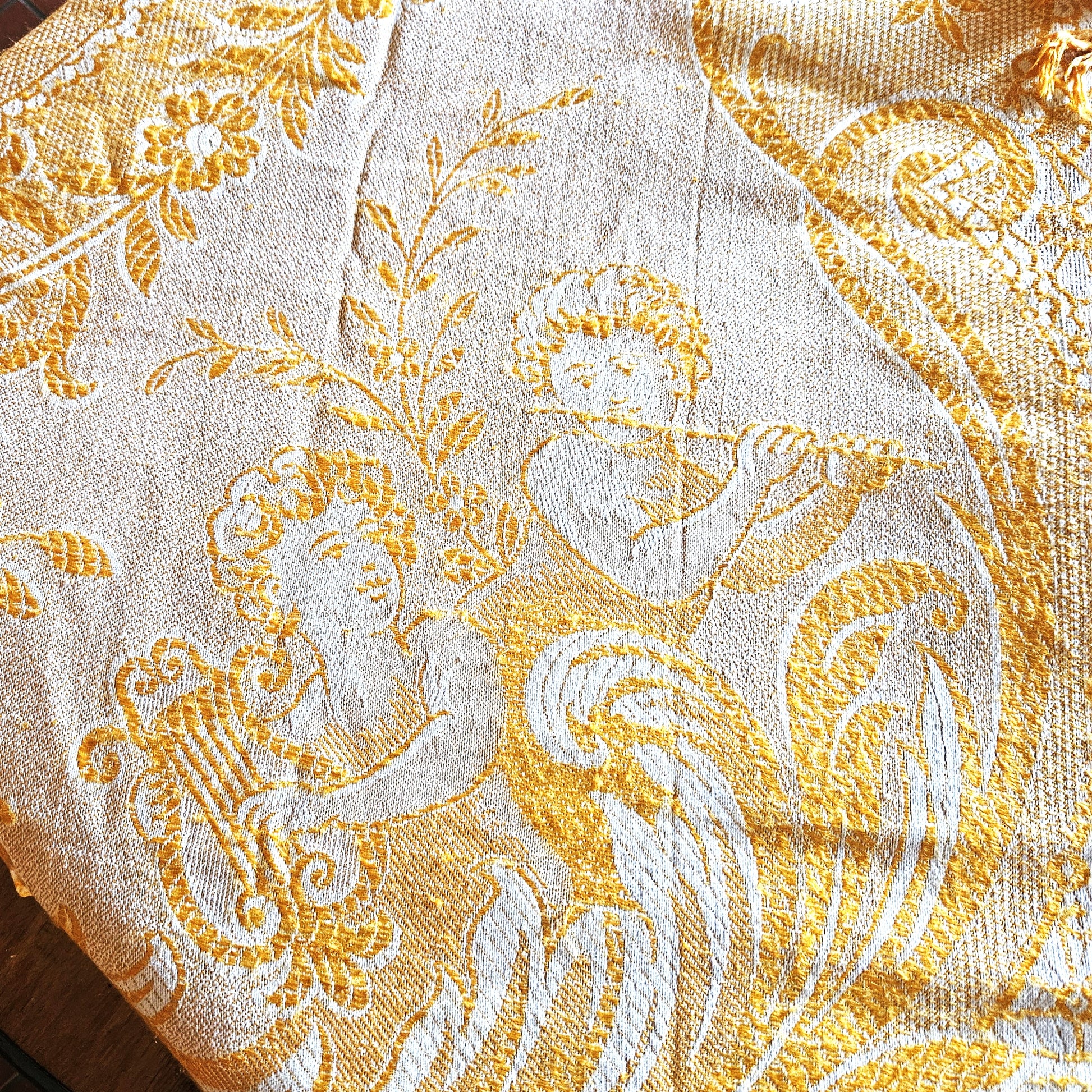 Détail d'un coin du jeté de canapé en jacquard double face années 1950-1960, montrant le motif figuratif d'un angelot musicien (putti) soufflant dans une trompette, entouré de rinceaux et feuillages dans un médaillon ovale, en camaïeu or sur fond crème, style Baroque/Louis XV.