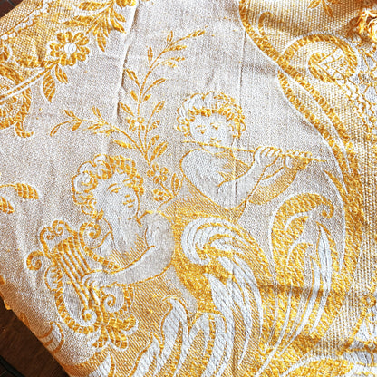 Détail d'un coin du jeté de canapé en jacquard double face années 1950-1960, montrant le motif figuratif d'un angelot musicien (putti) soufflant dans une trompette, entouré de rinceaux et feuillages dans un médaillon ovale, en camaïeu or sur fond crème, style Baroque/Louis XV.