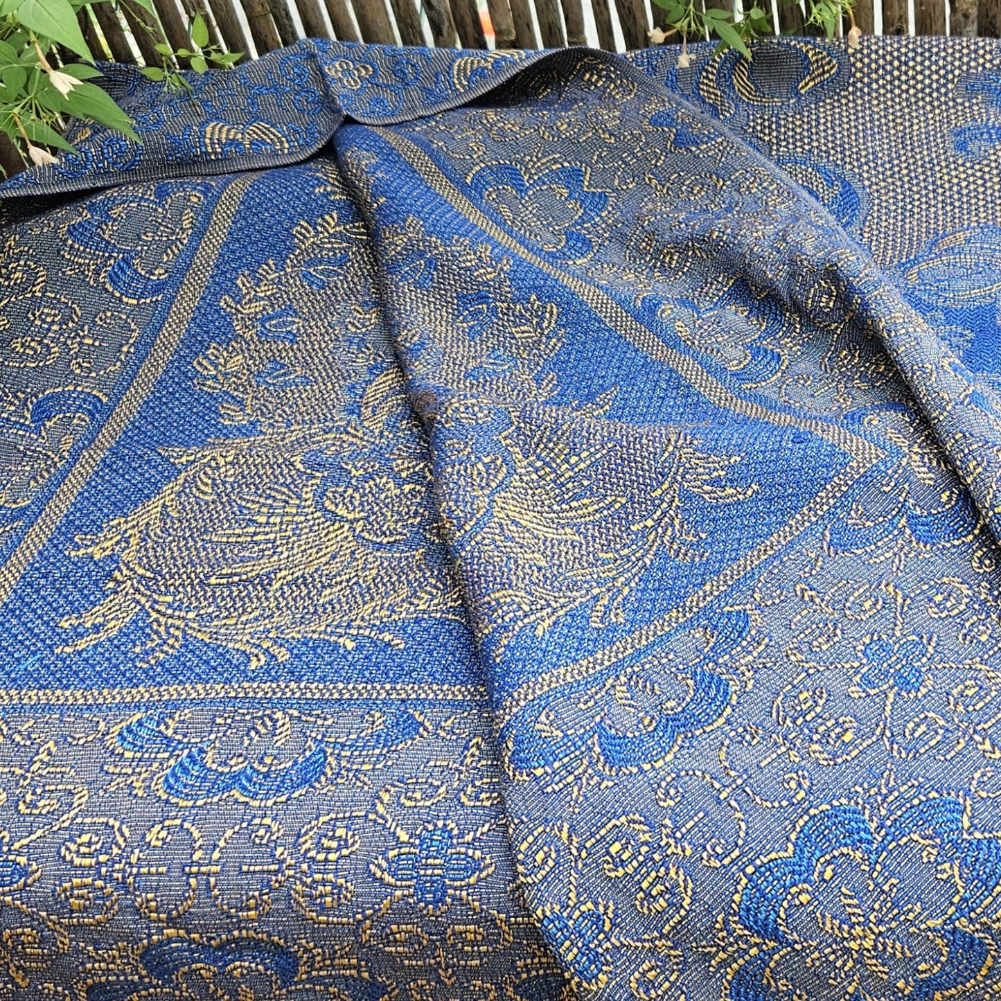 Vue générale drapée du jeté de canapé en jacquard double face années 1950-1960, posé sur un banc de jardin en bois, montrant les deux faces — bleu roi/jaune doré côté endroit et bleu marine/jaune ocre côté envers — avec la richesse du tissage jacquard et les multiples registres de motifs géométriques et floraux, fond de végétation.