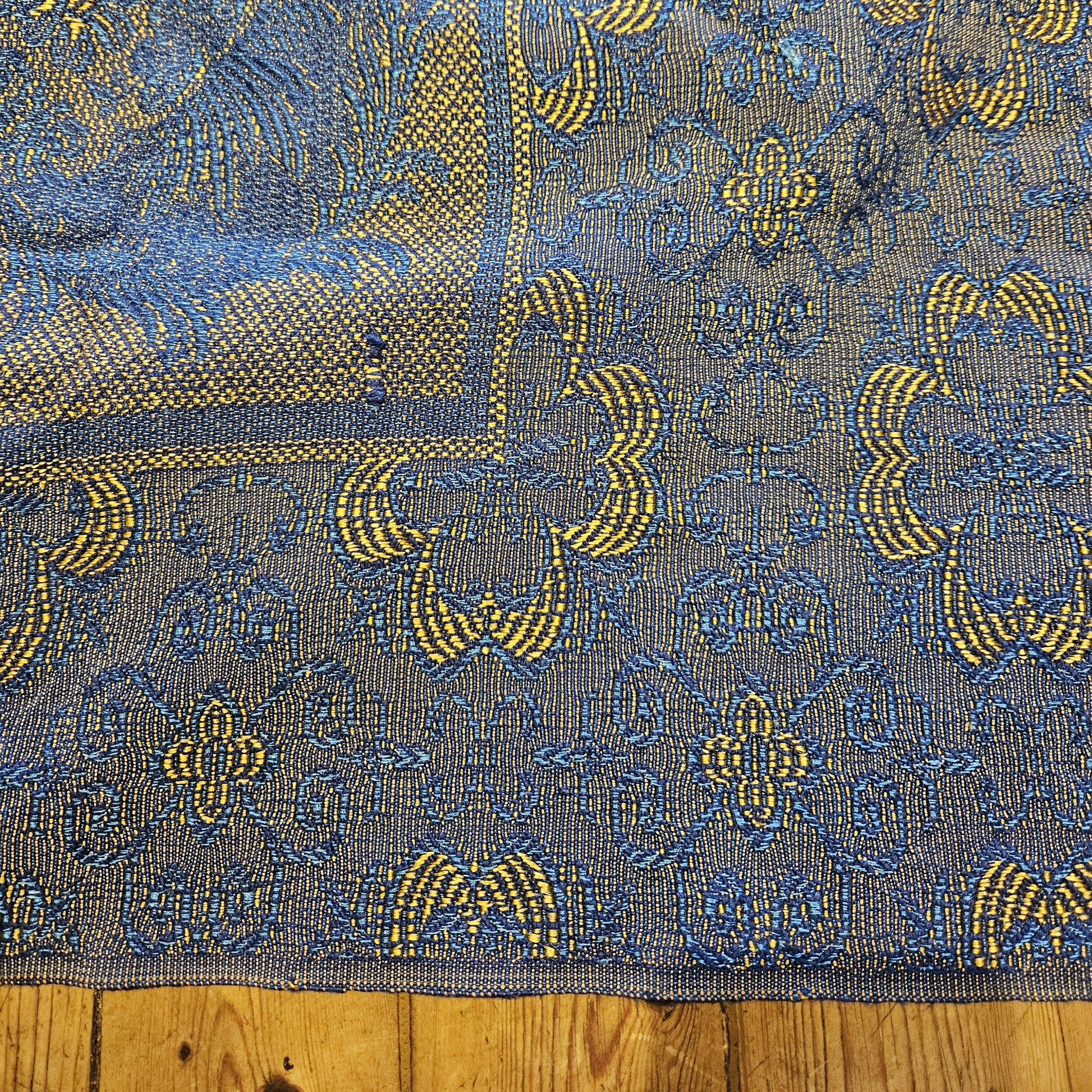 Détail de la bordure du jeté de canapé en jacquard double face années 1950-1960, montrant la frise à motifs géométriques en arceaux entrelacés jaune doré sur fond bleu marine, avec transition vers le registre central à rinceaux floraux, posé sur parquet en bois clair.
