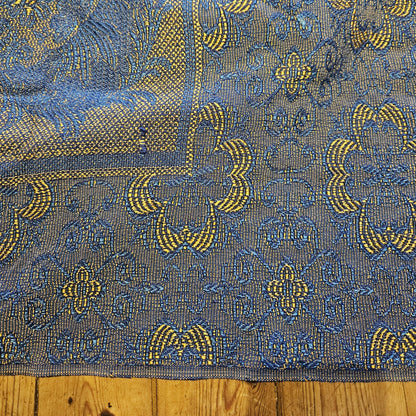 Détail de la bordure du jeté de canapé en jacquard double face années 1950-1960, montrant la frise à motifs géométriques en arceaux entrelacés jaune doré sur fond bleu marine, avec transition vers le registre central à rinceaux floraux, posé sur parquet en bois clair.
