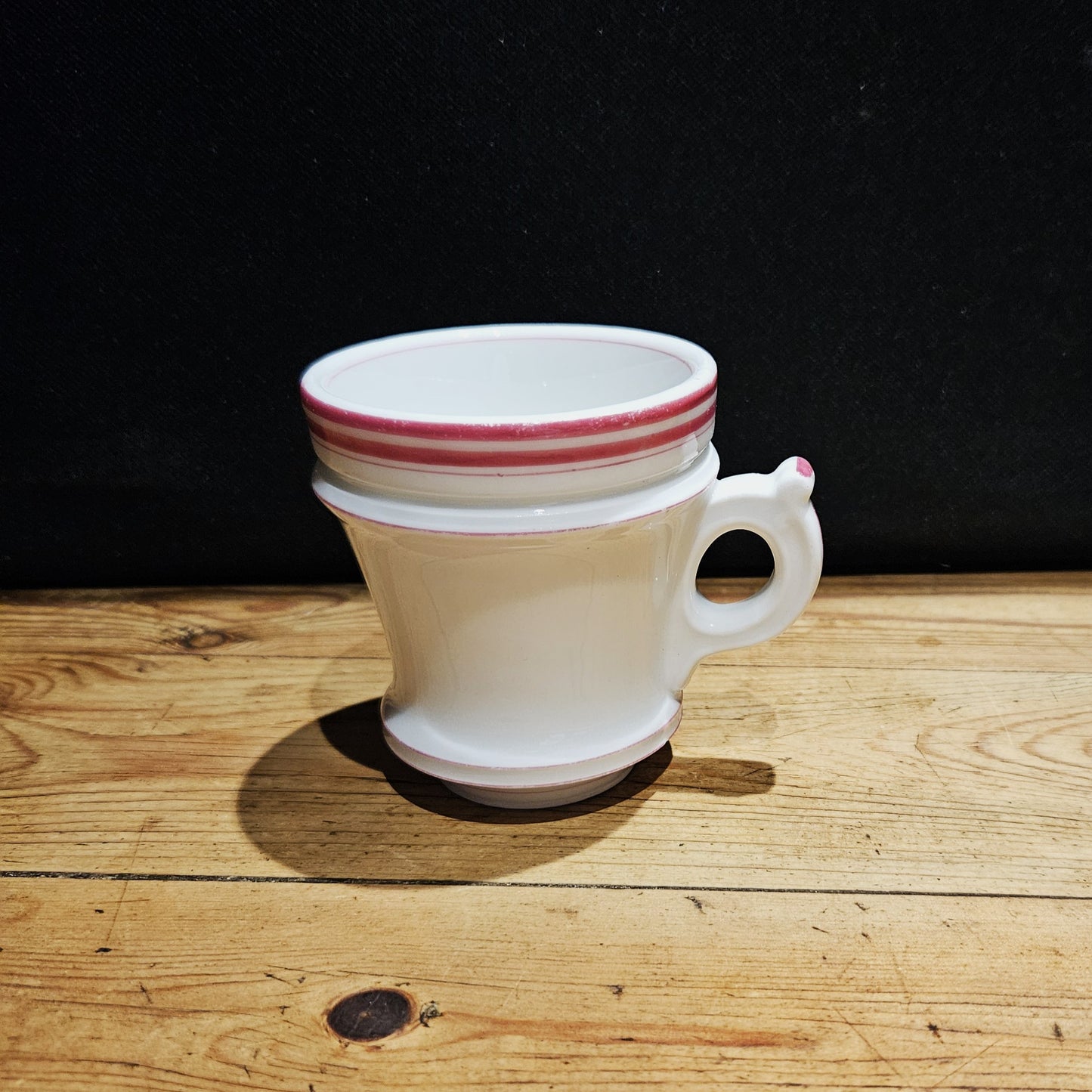 Tasse à brûlot ancienne en faïence blanche avec une petite anse ronde, ornée de trois rayures roses sur le bord supérieur, posée sur une surface en bois brut, fond noir.