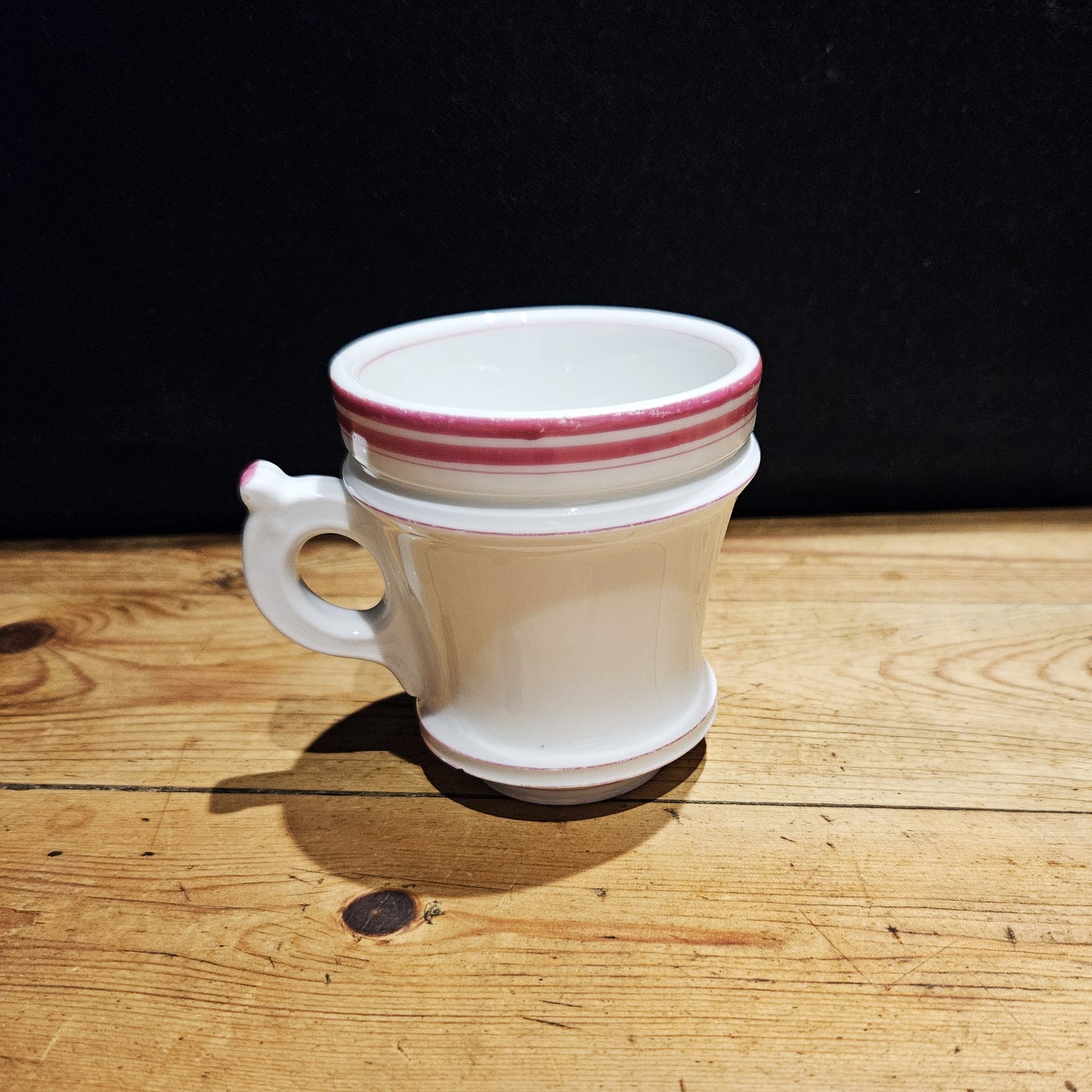 Tasse à brûlot ancienne en faïence blanche avec une petite anse ronde, ornée de trois rayures roses sur le bord supérieur, posée sur une surface en bois brut, fond noir.