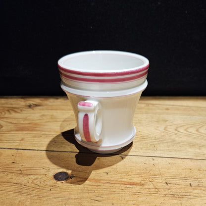 Vue de face de la tasse à brûlot en faïence blanche, avec trois rayures roses sur le bord supérieur, une petite anse rectangulaire ornée d'un filet rose, et un pied évasé, posée sur une surface en bois brut, fond noir.