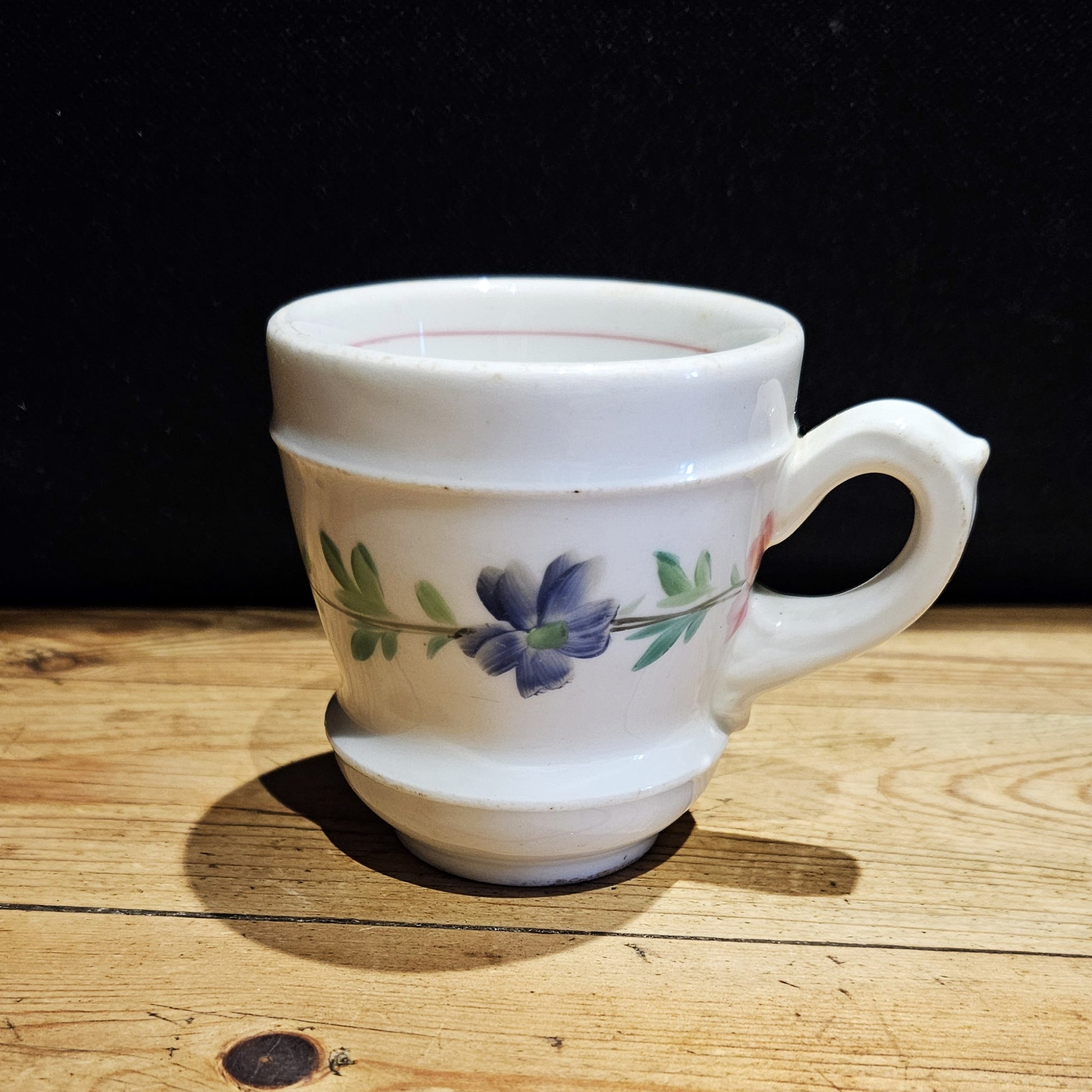 Tasse à brûlot ancienne en faïence blanche avec une petite anse en forme de larme, ornée d'une fleur bleue entourée de feuilles vertes  sur le milieu du corps de la tasse. Cette dernière est posée sur une surface en bois brut, fond noir.