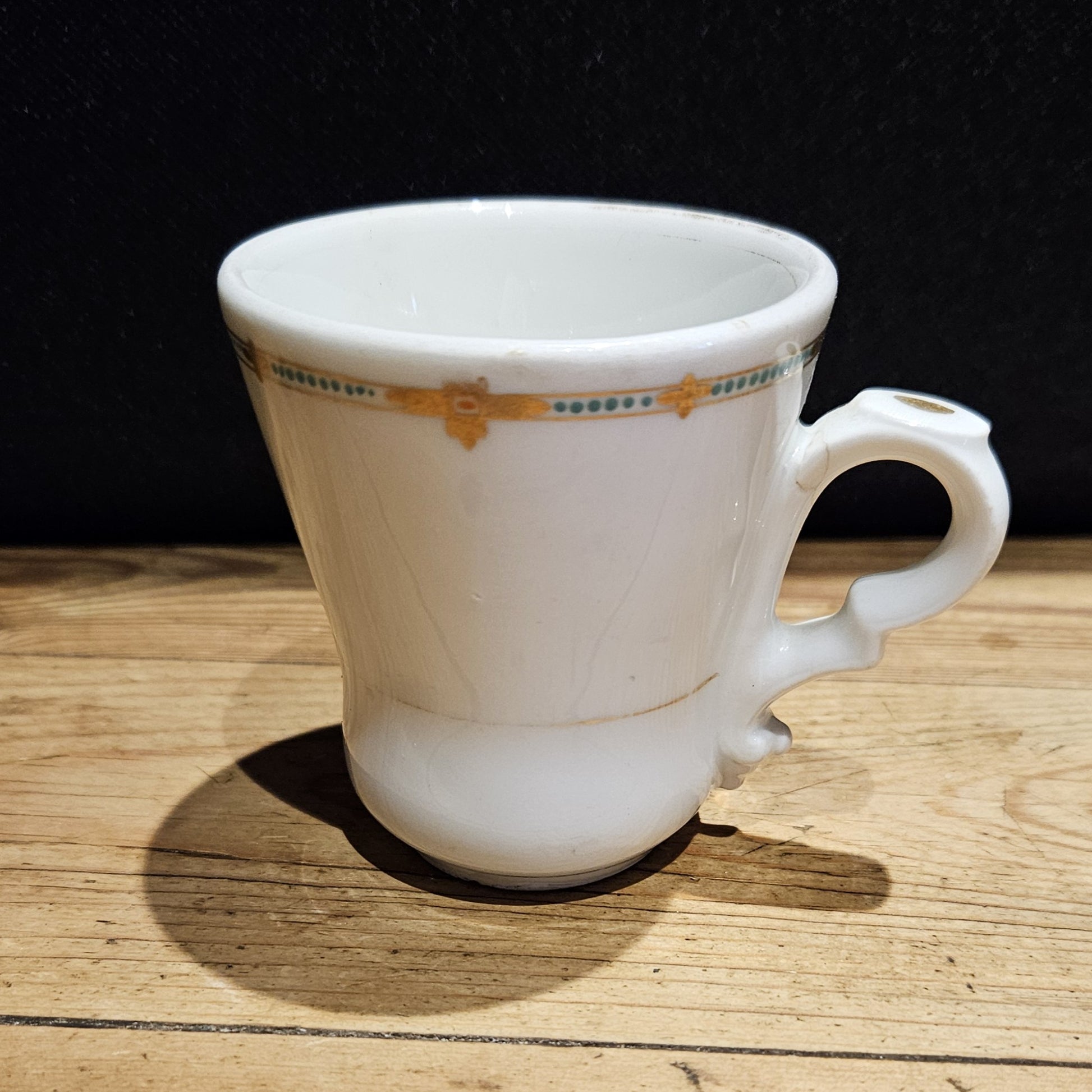 Tasse à brûlot ancienne en faïence blanche de style Art Déco, ornée d'une frise dorée et de points turquoise sur le bord supérieur, avec une anse en volute, posée sur une surface en bois brut, fond noir.