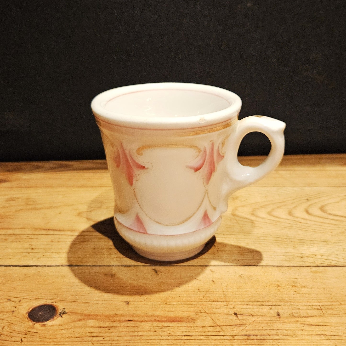 Tasse à brûlot ancienne en faïence blanche avec décor en relief de volutes et cartouches roses rehaussés de filets dorés, anse ronde, posée sur une surface en bois brut, fond noir.