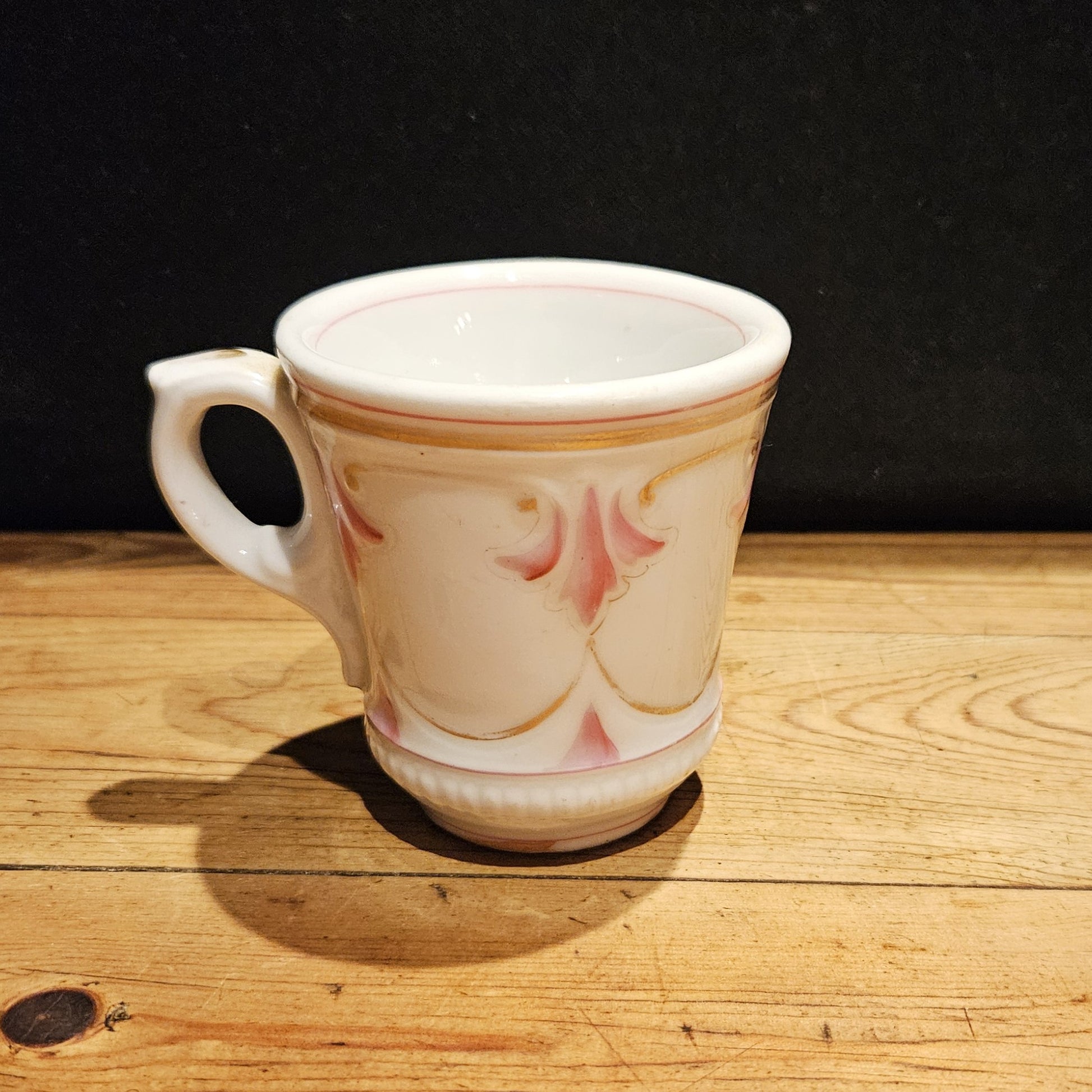 Vue de trois quarts de la tasse à brûlot en faïence blanche, mettant en valeur le décor en relief rose et doré et le pied annulaire, posée sur une surface en bois brut, fond noir.