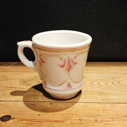 Vue de trois quarts de la tasse à brûlot en faïence blanche, mettant en valeur le décor en relief rose et doré et le pied annulaire, posée sur une surface en bois brut, fond noir.
